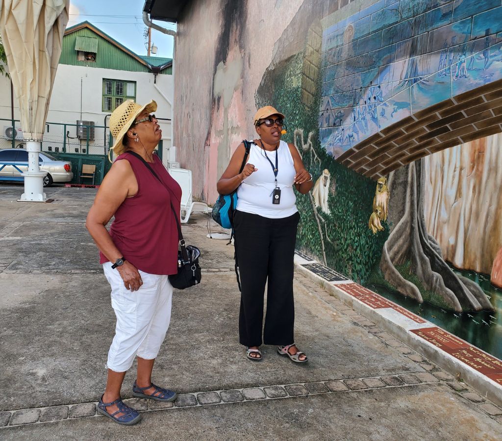 Tour guide Deborah enlightens a guest by Speightstown mural, unraveling its cultural tales.